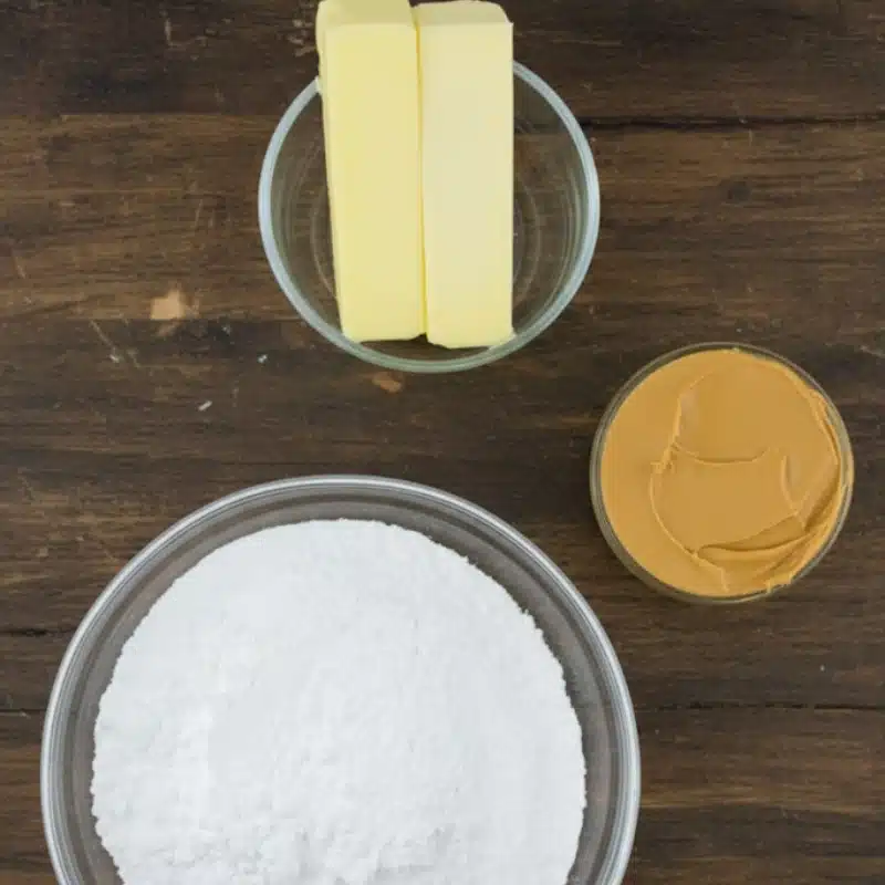 The three ingredients for peanut butter fudge—powdered sugar, butter, and creamy peanut butter—in separate glass bowls on a wooden table.