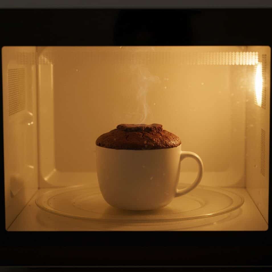 Mug cake baking inside a microwave with light on and cake rising.